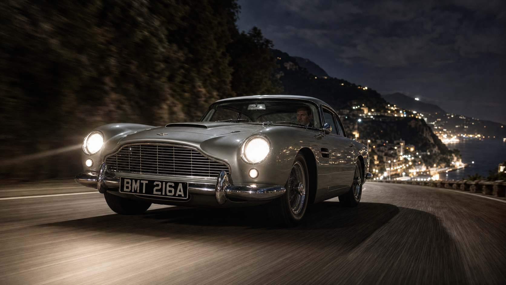 A Silver Birch Aston Martin DB5 in motion on a night-time European road, headlights blazing, cinematic low angle