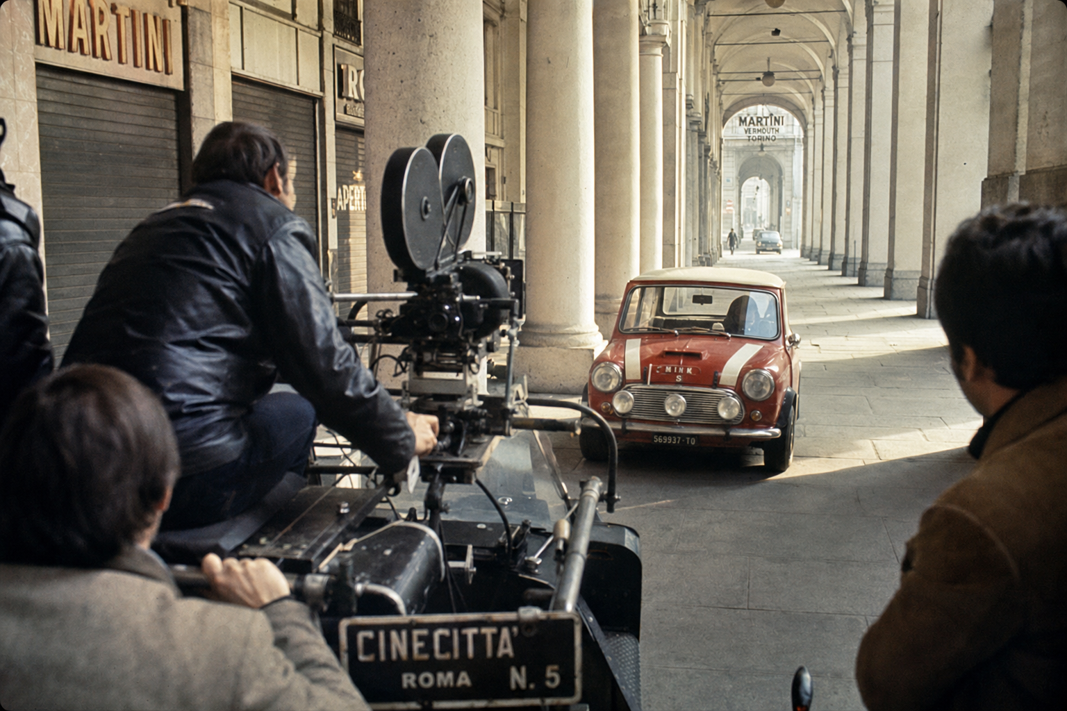 Film crew with camera mounted on vehicle filming a red Mini Cooper S through a Turin arcade